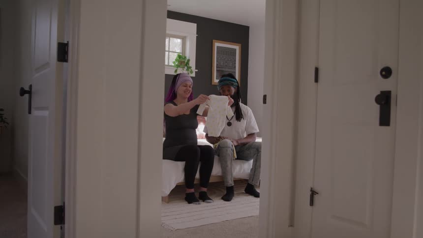 A happy pregnant woman and a man with dreadlocks are sitting on a bed in a bedroom and looking at a white baby onesie