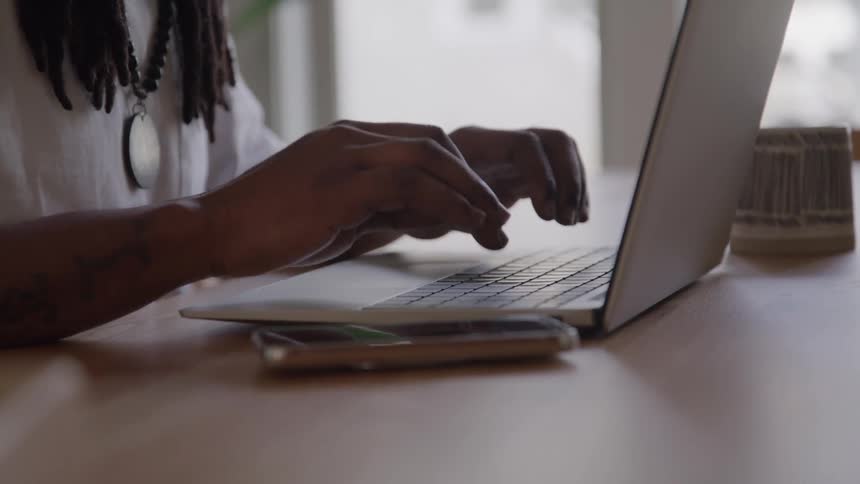 A close view of a mans hands with tattoos on his arm shows him typing on a laptop computer with a phone lying next to it on a wooden desk