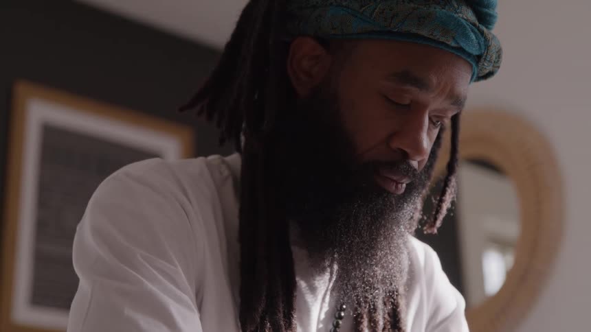 A man with dreadlocks and a long beard looks down with a calm expression with a faint smile on his face in a bright home setting