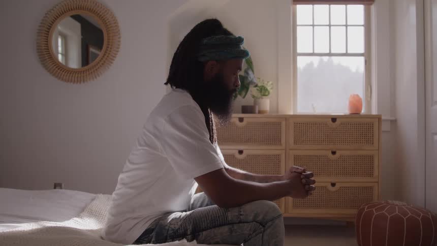 A man with a beard and dreadlocks is sitting on a bed in a sunlit bedroom with his hands clasped together