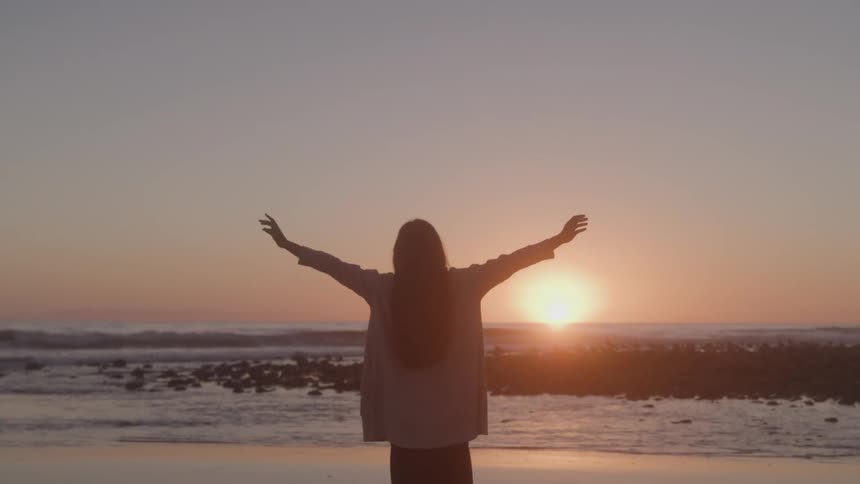 A woman stands on a rocky beach at sunset facing the sun with her arms raised in the air