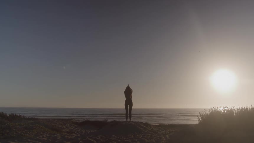 A slender woman in silhouette stands on a sandy beach and performs a yoga pose with her arms raised above her head facing the ocean and the sun setting on the horizon