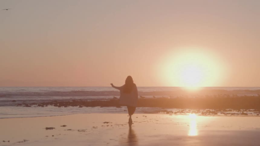 A silhouette of a woman walks barefoot on a wet beach at sunset as she walks toward the gentle ocean waves