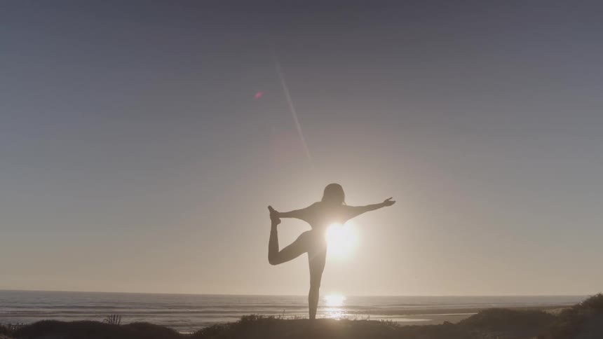 Woman at sunset on the beach lifting one leg to shoulder height and holding it with one arm while the other arm is stretched out to the side for balance