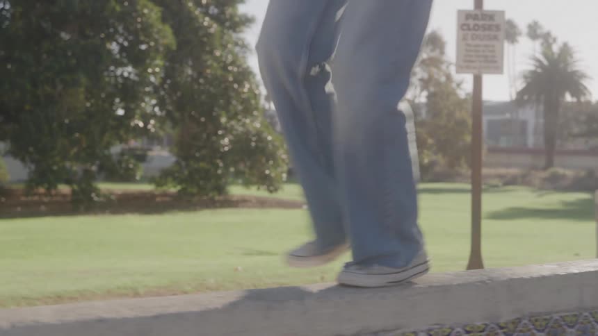 A lowangle tracking shot follows a person in wideleg jeans and sneakers as they walk and balance along the top of a decorative tile wall in a park