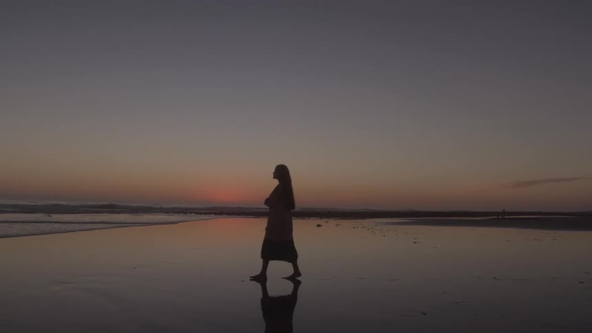 Woman walking barefoot on wet beach sand toward the ocean at sundown
