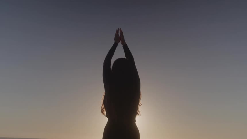 Woman doing yoga on the beach raising both hands in the air facing the orange setting sun