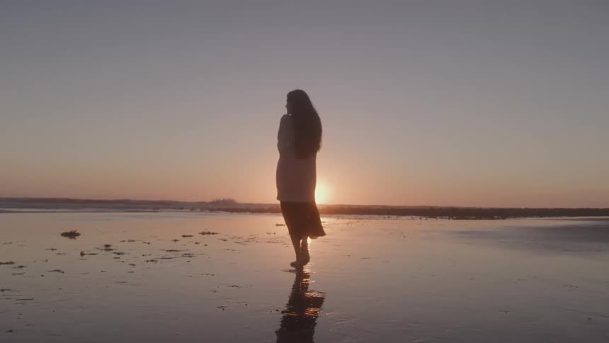 Woman walks barefoot on the beach during sunset