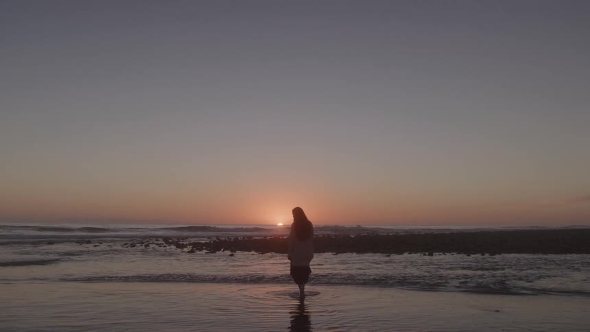 Woman walking on shallow water on the beach with the orange setting sun in the distance