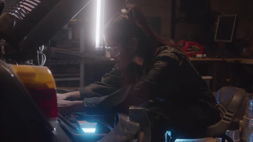 Woman in overalls works focused on a vehicle engine inside the workshop