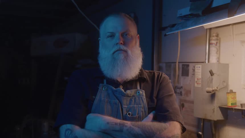 Man in an apron crosses his arms leaning against a workshop bench
