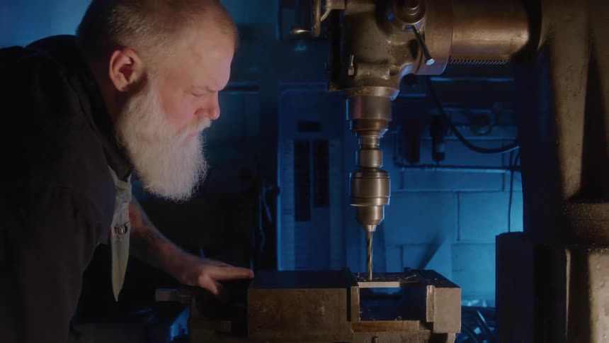 Man with a white beard operates a drill press in the workshop