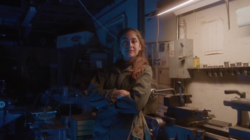Woman in overalls crosses her arms leaning against a workshop bench