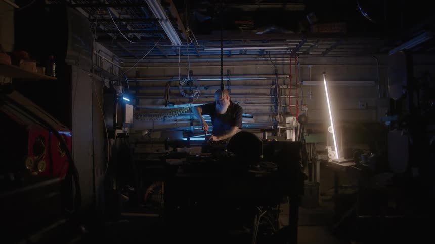Man with a white beard works at a lit bench inside the workshop