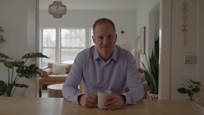 Man in a dress shirt holds a mug while sitting at the kitchen table