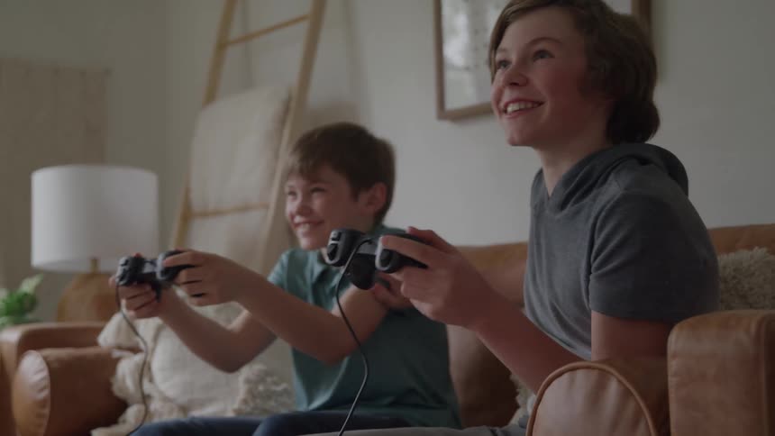 Two boys sitting on a couch playing video games excitedly as the teenage boy playfully shoves the younger kid with one hand making them both laugh