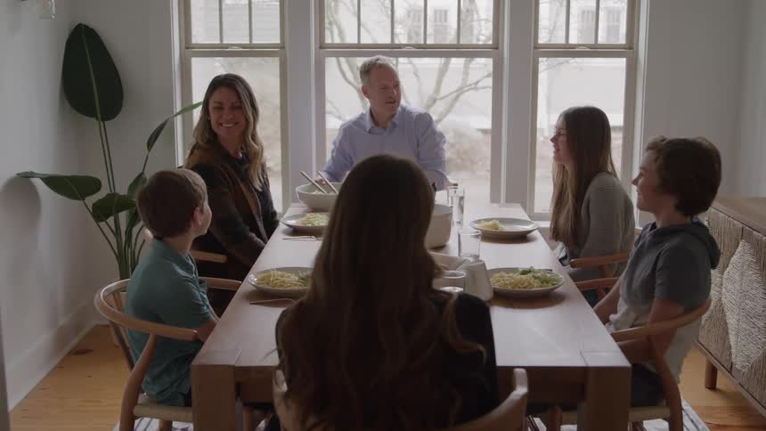 Family of six sitting together at the dining table for breakfast smiling warmly a father and mother with two young boys and two teenage girls enjoying the meal
