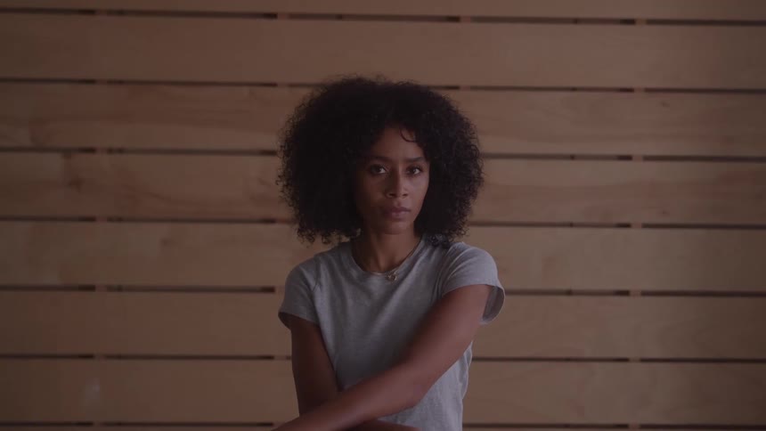 Young woman with long curly hair standing indoors beside a wooden wall looking sad