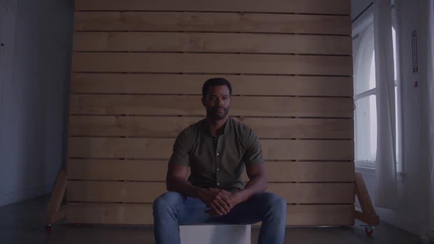 A man with a beard sits on a white cube in a room with a wooden slatted wall and a large window