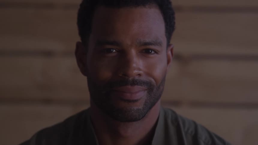 A smiling man with a beard is looking directly at the camera with a wooden wall and soft lighting in the background
