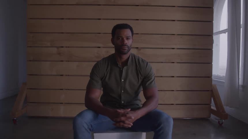 A shot of a handsome black man sitting on a white box in front of a wooden slatted wall he is wearing a dark green shirt and jeans and looks straight into the camera