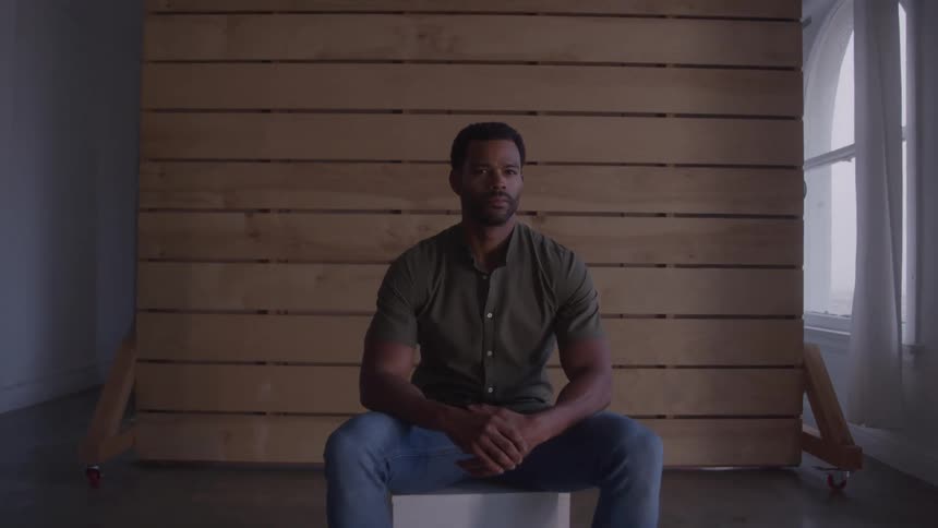 A man sits indoors on a cube seat in front of a wooden backdrop