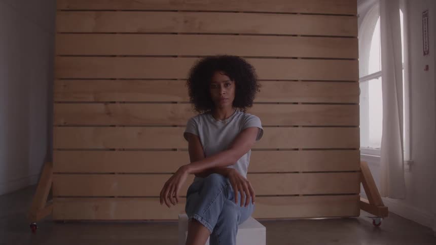 A woman with curly hair sits on a white stool with her arms crossed and resting on her knees in front of a wall made of wooden slats