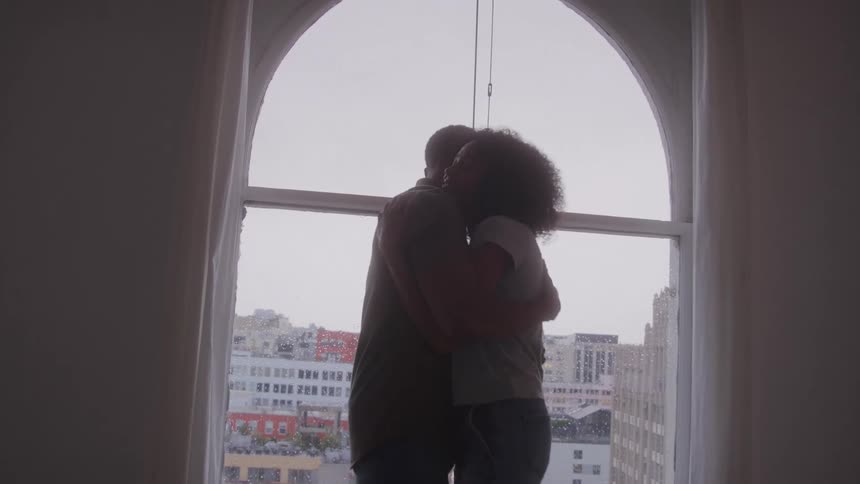 A video shows the silhouette of a couple standing in front of a large arched window the man pulls the woman into a tight embrace as they look out at the cityscape below