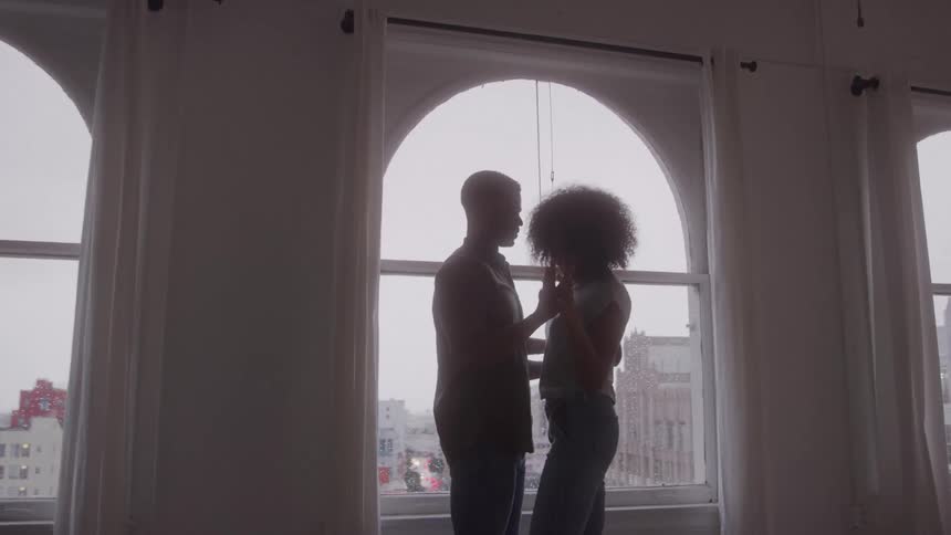 A couple is seen in silhouette from the waist up facing each other in front of a window with a city view they are holding each other and appear to be dancing or swaying gently