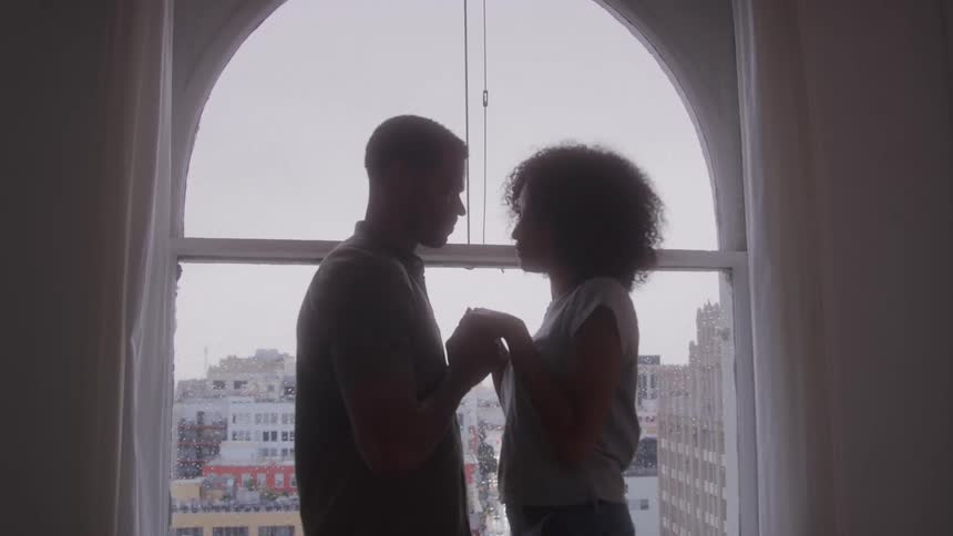 A silhouette shot shows a couple standing close together holding hands in front of a large arched window the city skyline is visible in the background through the window