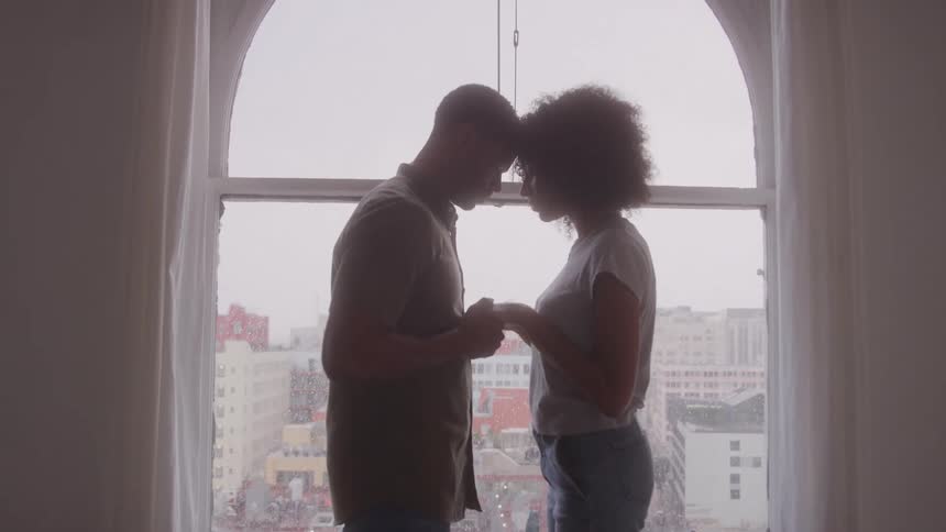 A man and a woman standing hand in hand their faces touching in front of a window overlooking the city
