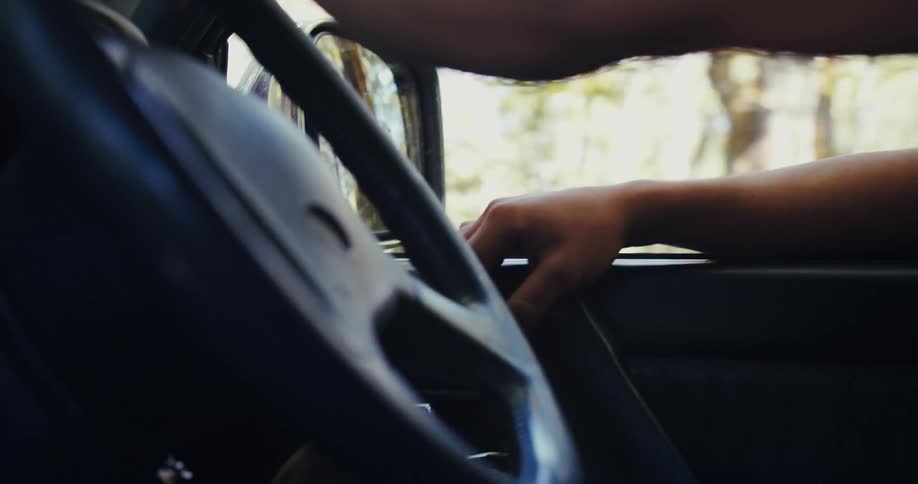 A mans hand holding the steering wheel of a car with his other arm resting on the car door