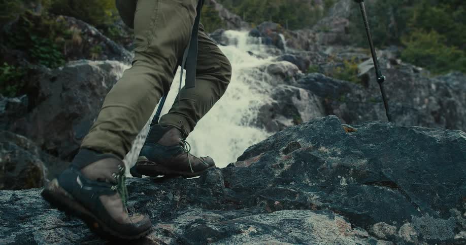 Person holding a guiding stick walking up a rocky terrain at midday as water flows down the rocks