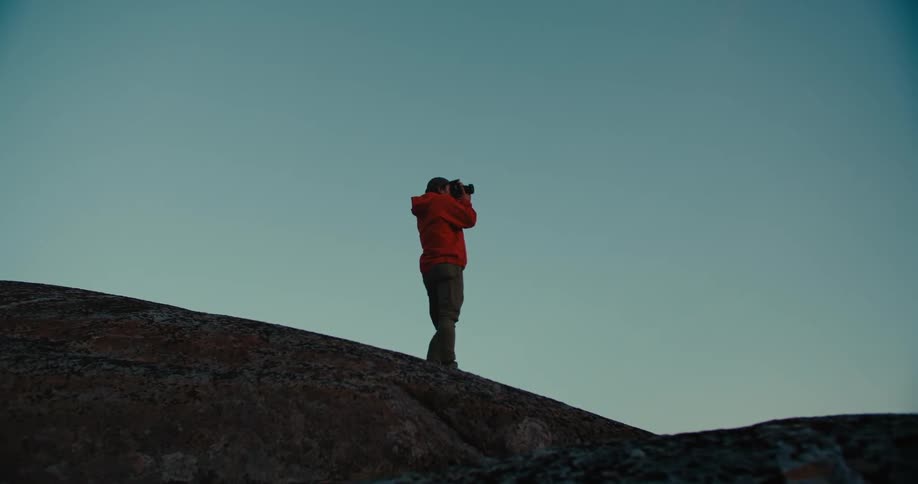 A person standing on a large boulder using a professional camera to take a photograph
