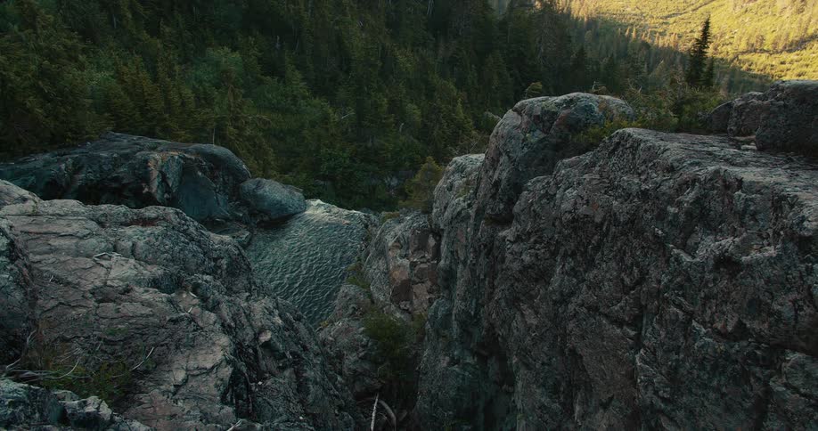 A scenic shot captures a lush green forest on a distant mountain slope with a rocky cliff face in the foreground a small cascading waterfall is visible between the large boulders