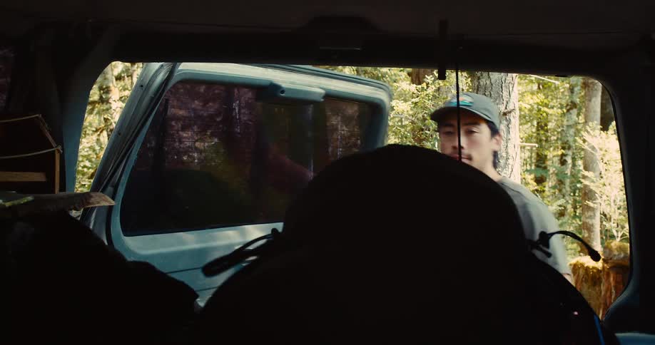 A person is shown from inside a dark vehicle looking out the back window the back of another vehicle and a forest are visible outside a man in a baseball cap appears and begins to pull a backpack out of the vehicles rear