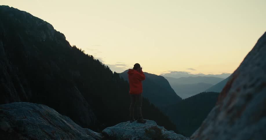 A photographer in an orange jacket stands on a rocky mountain summit at sunrise taking a picture of the vast misty valley below