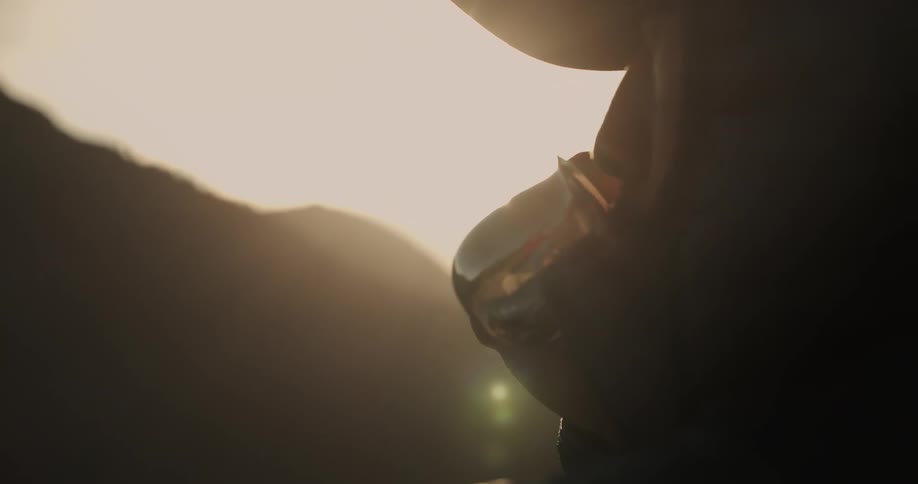 A man wearing a hat with protective wings over the ears drinks from a metal mug in front of a mountain