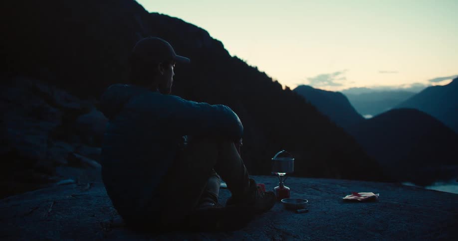 A man is sitting on a rock in a mountainous landscape at dawn with a camping stove and equipment next to him