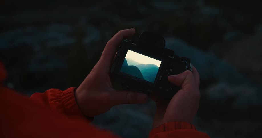 A closeup shot of a persons hands holding a black camera they are wearing an orange jacket and appear to be taking a picture of something in the distance the screen on the camera shows a shot of a mountain range