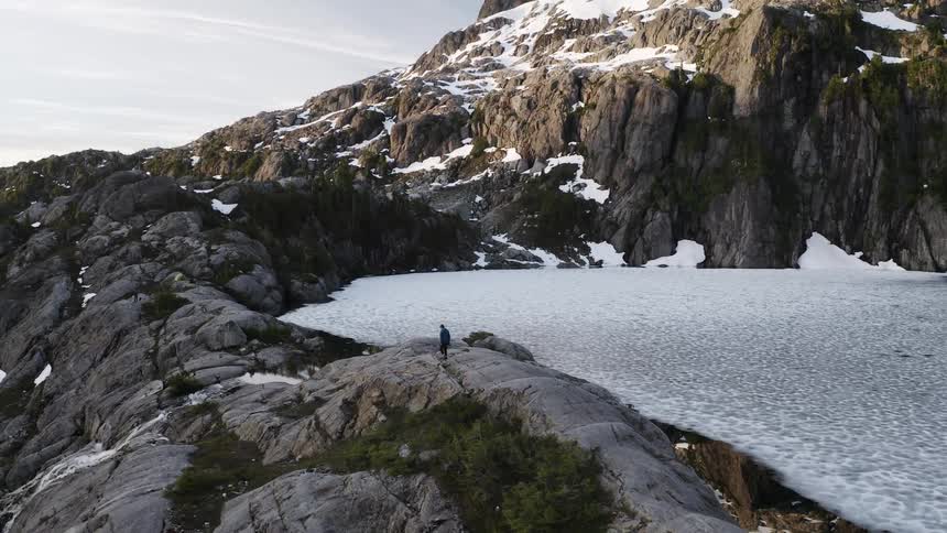 A person walks beside a frozen lake with snow capped mountains