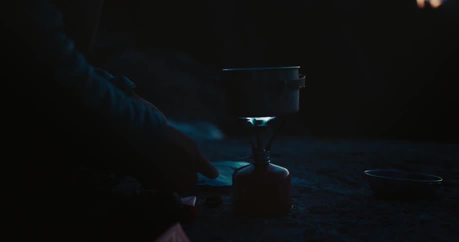 A person ignites a portable stove to heat a pot during nighttime outdoors