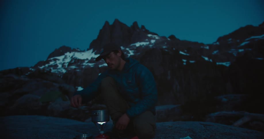 A man is crouching in front of a small camping stove and cooking on a rocky surface with a massive snowcovered mountain in the background at night