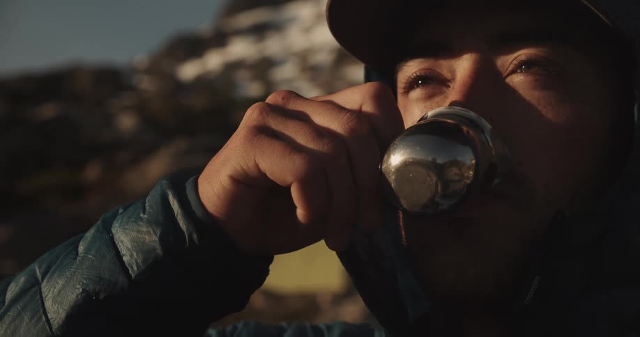 A person drinks from a metal cup at sunset with mountains in the background