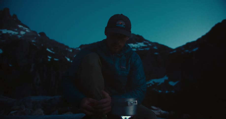 Man sitting prepares food on a stove at dusk in the mountains