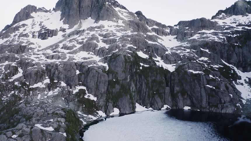 A dramatic mountain range with snowcovered peaks and jagged rock faces surrounds a large icy lake in a wild remote landscape