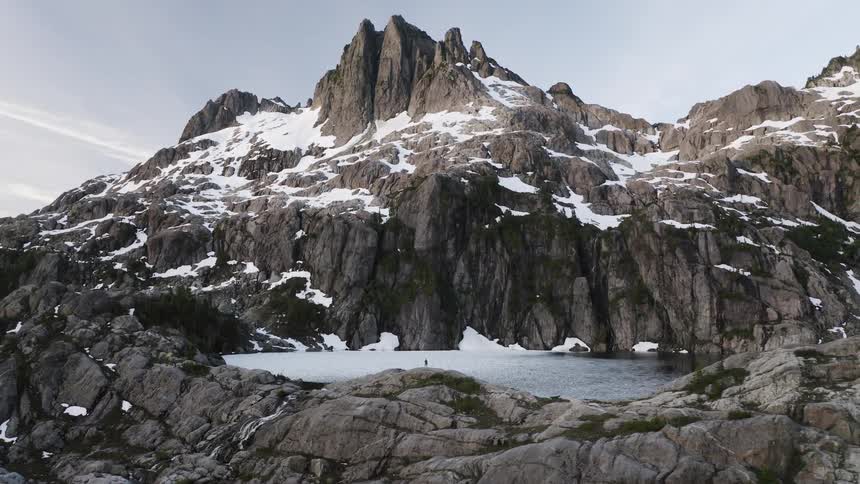 A person stands by a frozen alpine lake beneath snow covered mountains