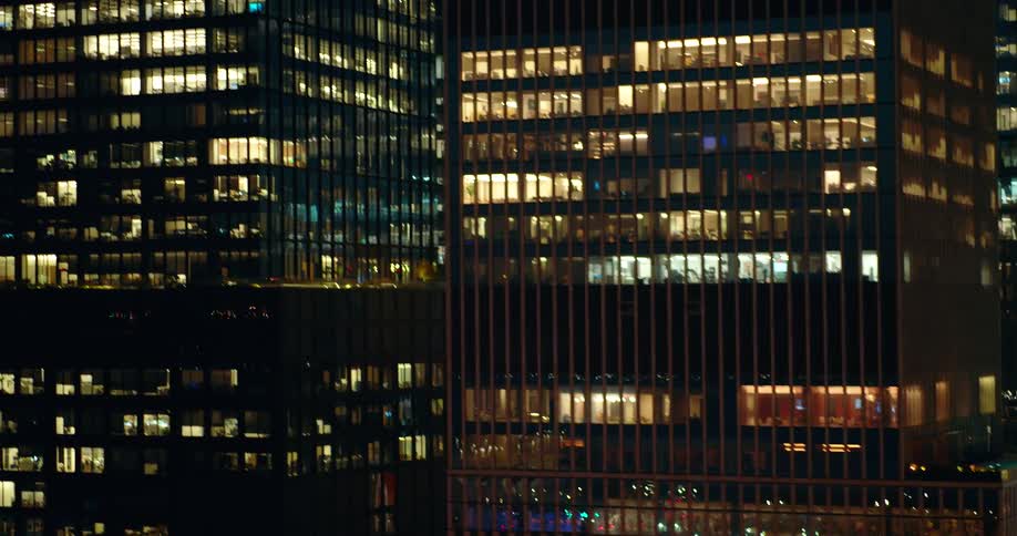 A wide dark view of a modern highrise office building at night shows numerous floors with lights on in the windows revealing a glimpse of the interiors