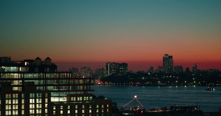 A city skyline with glowing lights reflects over a river at sunset