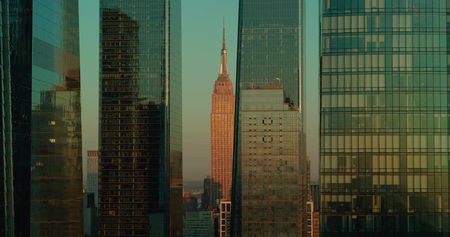 The empire state building glows warmly at sunset framed tightly between towering glass skyscrapers